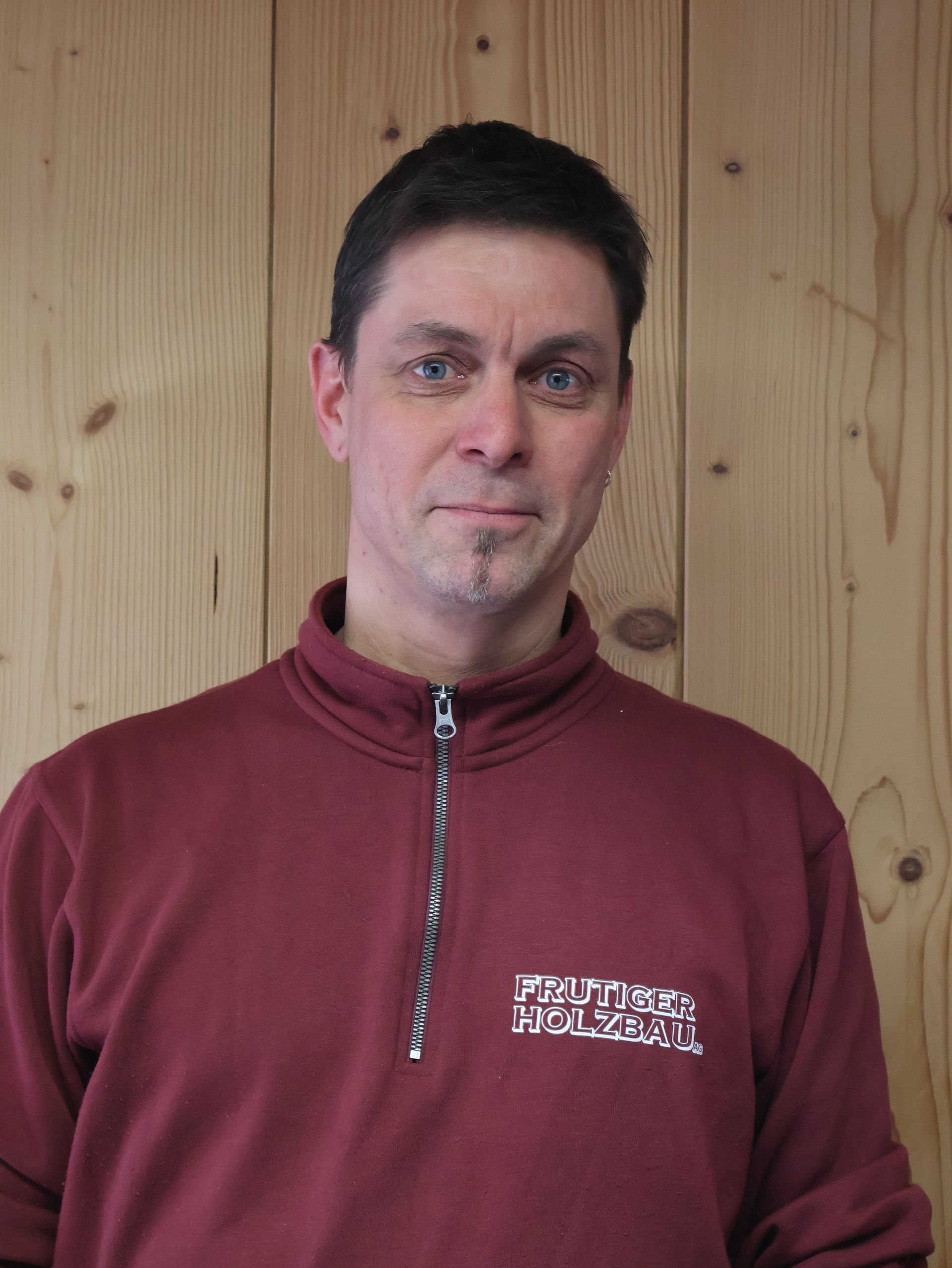 Ein Mann lächelt in die Kamera; er trägt ein dunkles T-Shirt. Im Hintergrund ist eine weisse Wand zu sehen.
