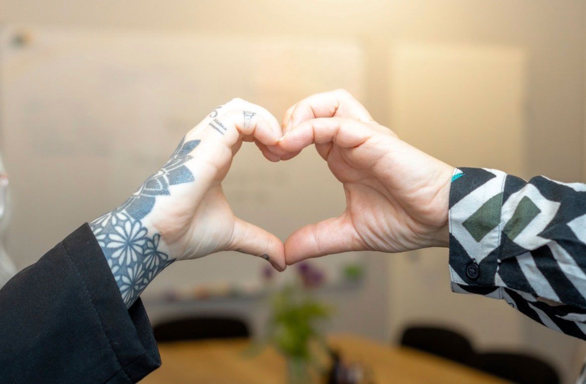 Zwei Hände, die ein Herz formen, eine davon mit einem Tattoo, vor einem verschwommenen Innenraumhintergrund