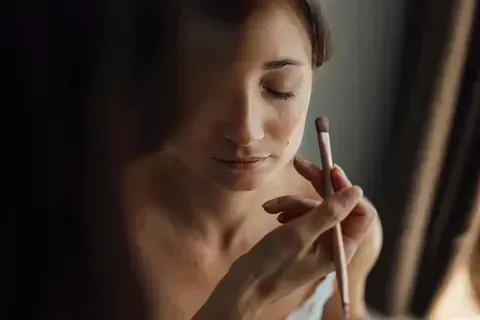 Mujer a la que le aplican maquillaje con brocha cerca de una ventana.