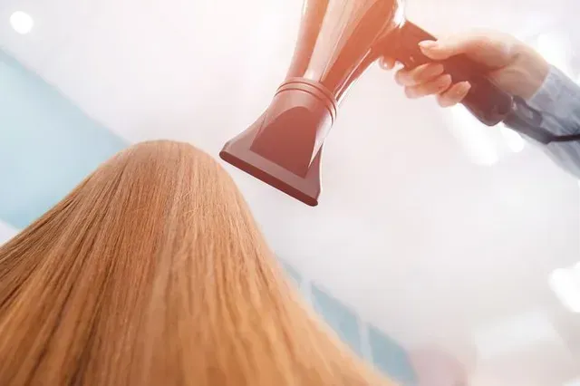 Peluquera secando cabello largo, castaño claro con un secador negro en un salón de belleza.