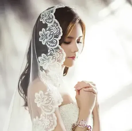 Mujer con velo de encaje blanco, manos entrelazadas, vestida con un vestido blanco, al aire libre.