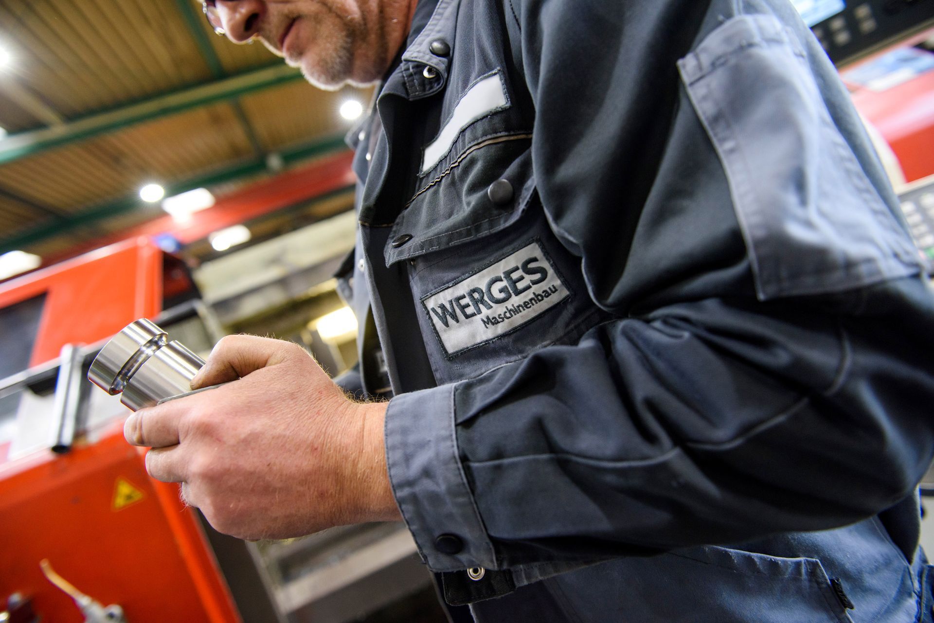 Ein Arbeiter in blauer Arbeitskleidung hält ein Metallteil in der Hand, Fabrikszene. Das „WERGES“-Logo ist sichtbar.