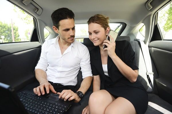 Um homem digitando em um laptop e uma pessoa falando ao telefone estão sentados juntos no banco de trás de um carro.