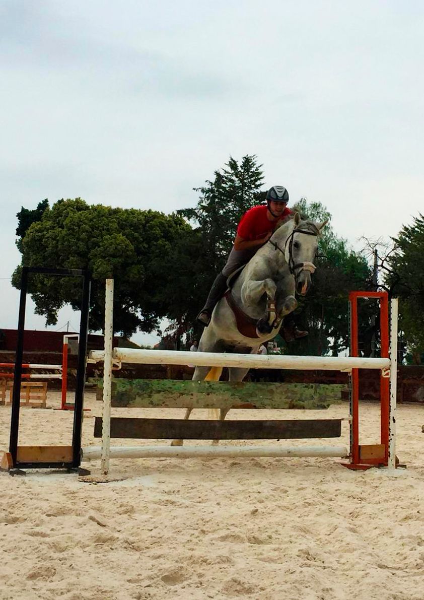 Jinete sobre un caballo gris saltando un obstáculo en una pista ecuestre arenosa al aire libre.