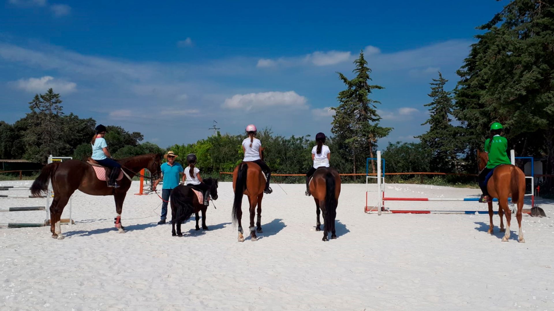 Gente montando a caballo en una pista al aire libre, con obstáculos para saltar y árboles bajo un cielo azul.