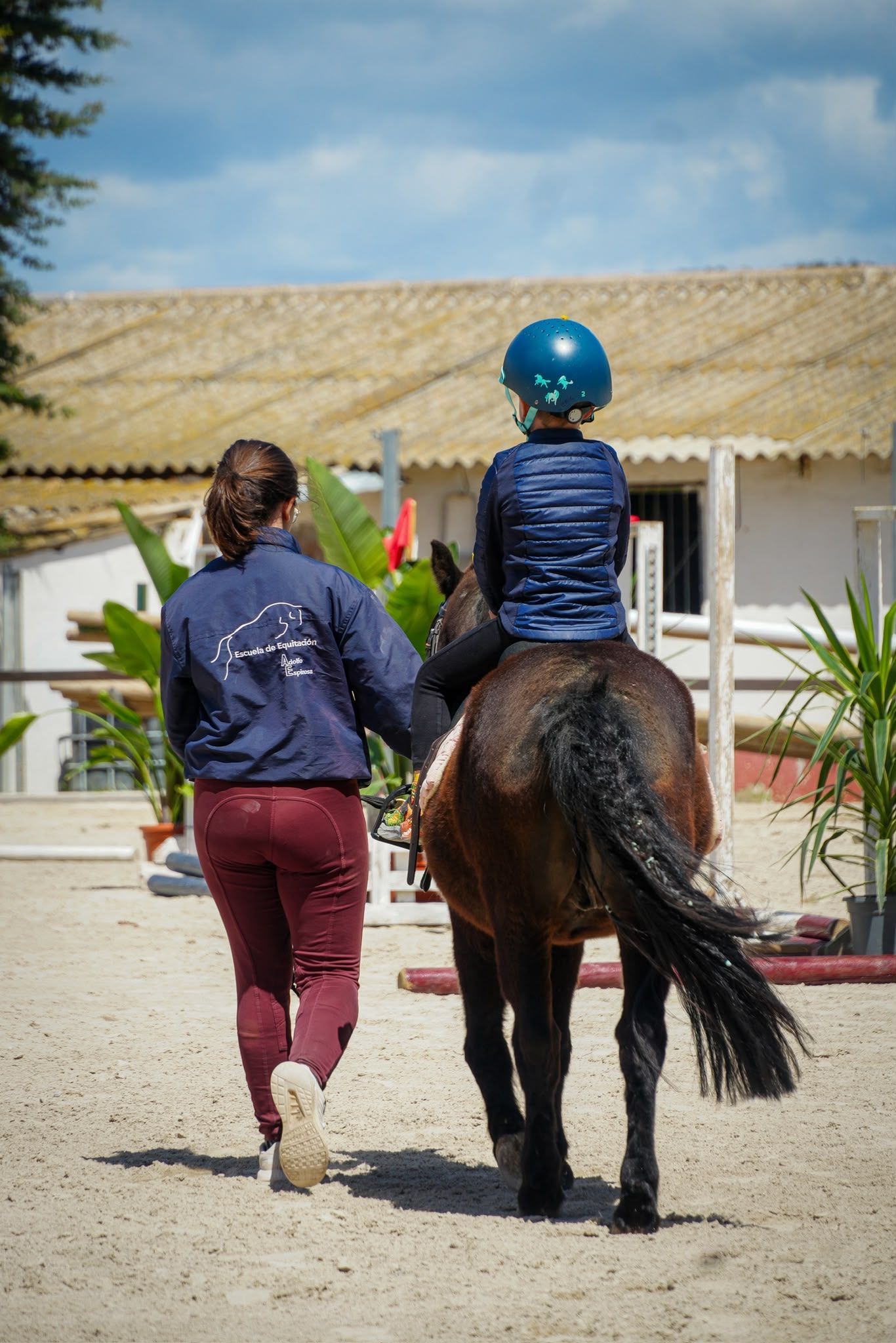 Persona que guía un caballo con un niño a caballo, vista desde atrás en una pista de equitación al aire libre.