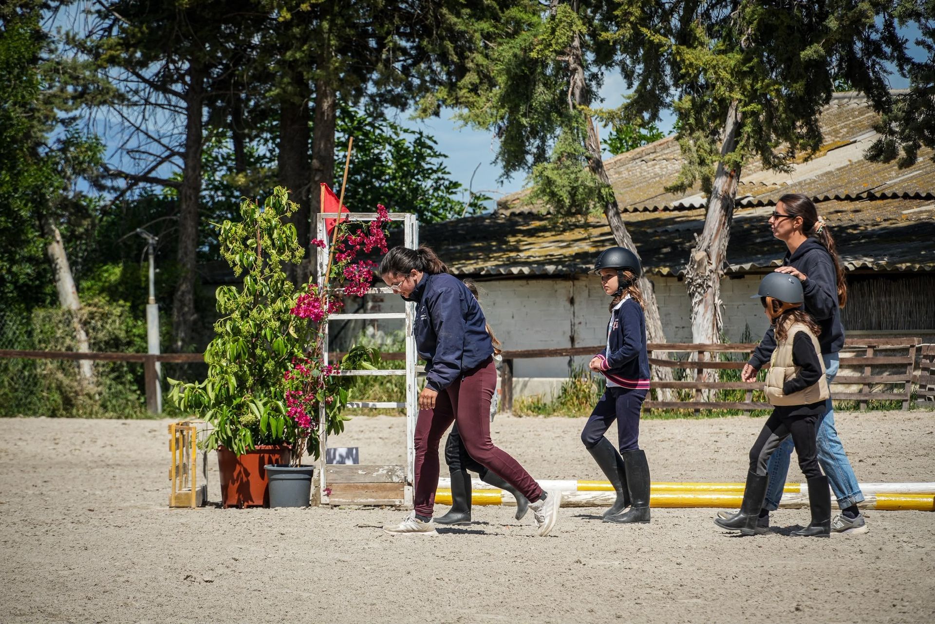 Tres personas pasan junto a macetas con flores y una rampa de salto en una soleada pista al aire libre.