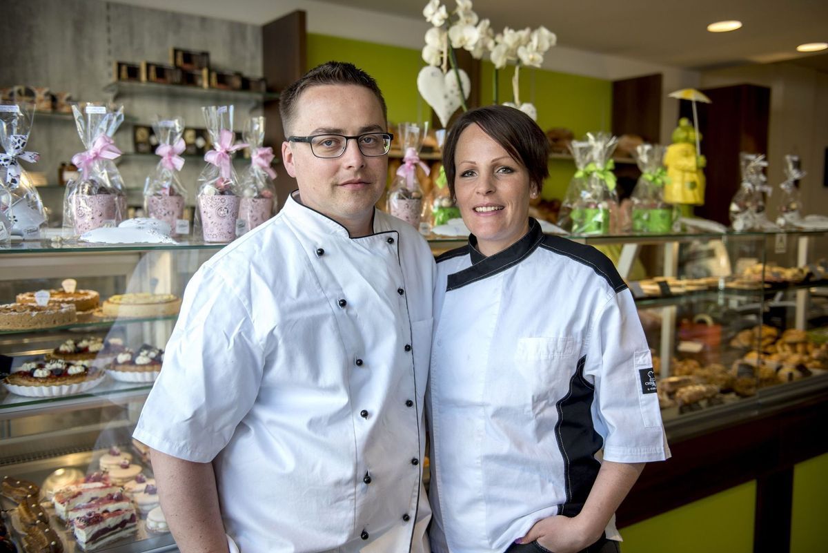 Deux boulangers en blouse blanche souriant dans une boulangerie, entourés de pâtisseries.