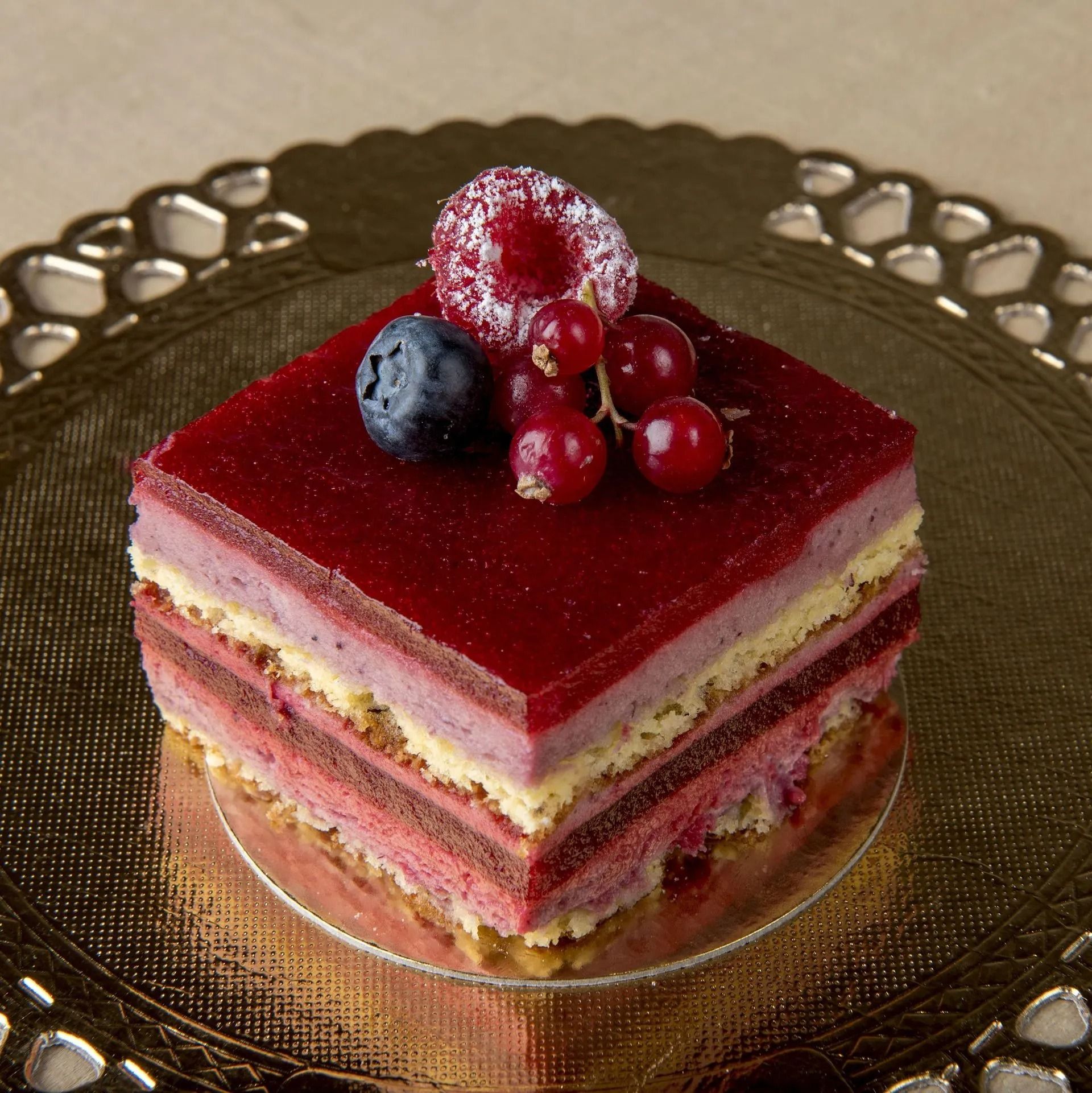 Gâteau aux baies carré sur une assiette décorative dorée, garni de baies rouges, de myrtilles et de sucre en poudre.