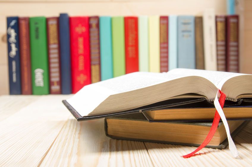 Libro abierto con marcapáginas de cinta roja sobre una mesa de madera, libros coloridos difuminados al fondo.