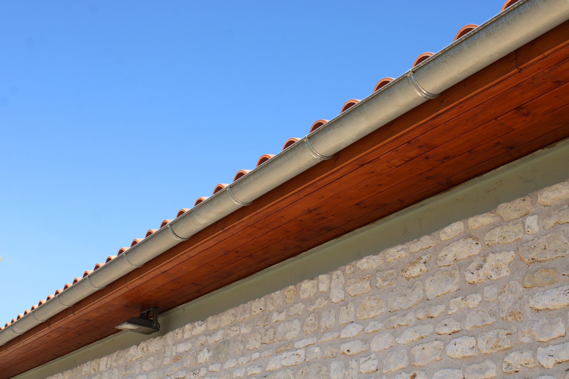 Mur d'une maison en pierre avec avant-toit en bois et toit de tuiles rouges se détachant sur un ciel bleu.