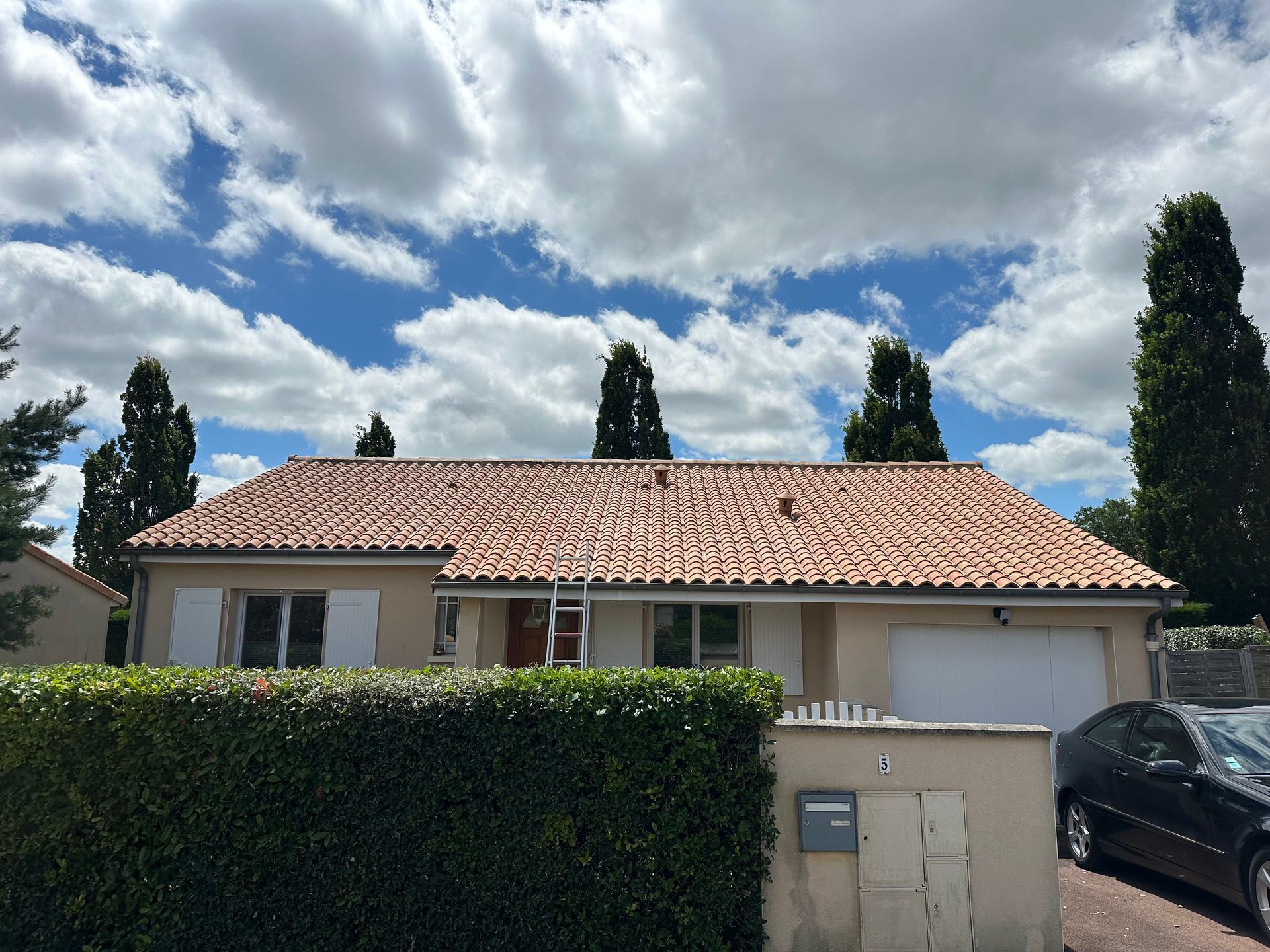 Maison de banlieue avec toit en tuiles de terre cuite, murs blancs, haie et voiture garée le long d'une allée sous un ciel nuageux.