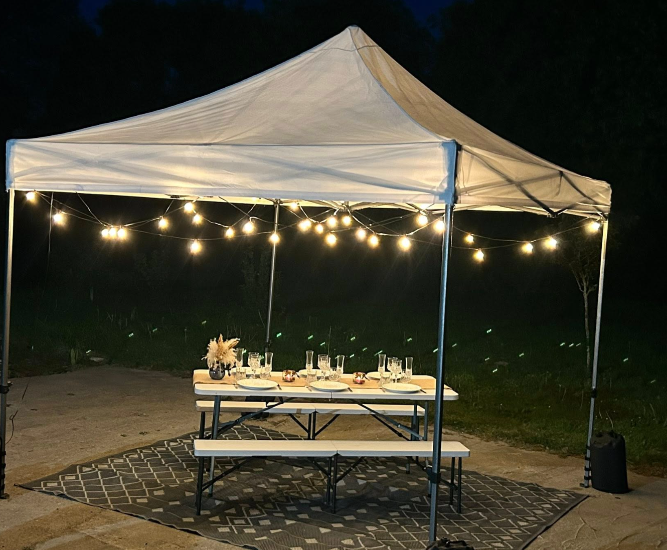 Tente blanche avec guirlandes lumineuses au-dessus d'une table de pique-nique pour un repas en plein air le soir.