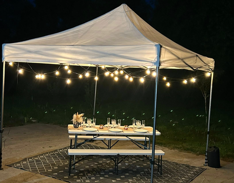 Longue table dressée pour un rassemblement sous une tente, décorée de verdure, de lumières et de couverts.