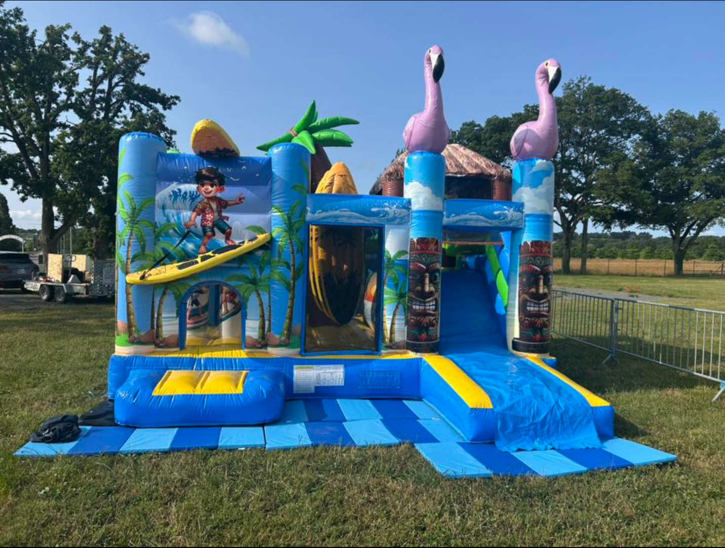 Maison gonflable à thème tropical avec toboggan, palmiers et flamants roses sur terrain en herbe.