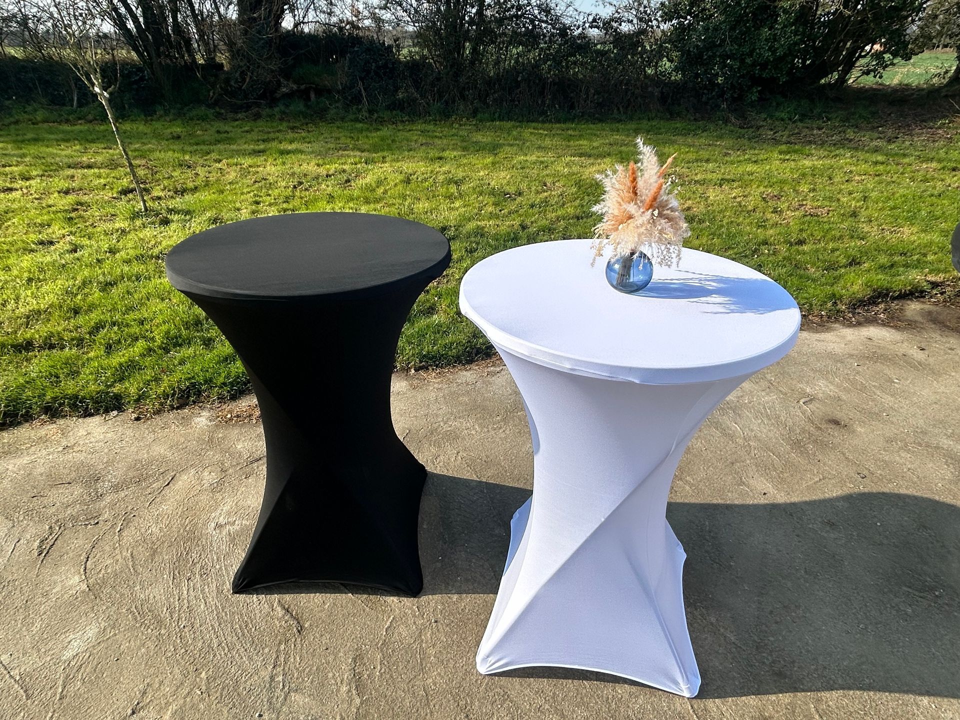 Deux tables basses, une noire et une blanche, en extérieur. La blanche est ornée d'une petite composition florale.