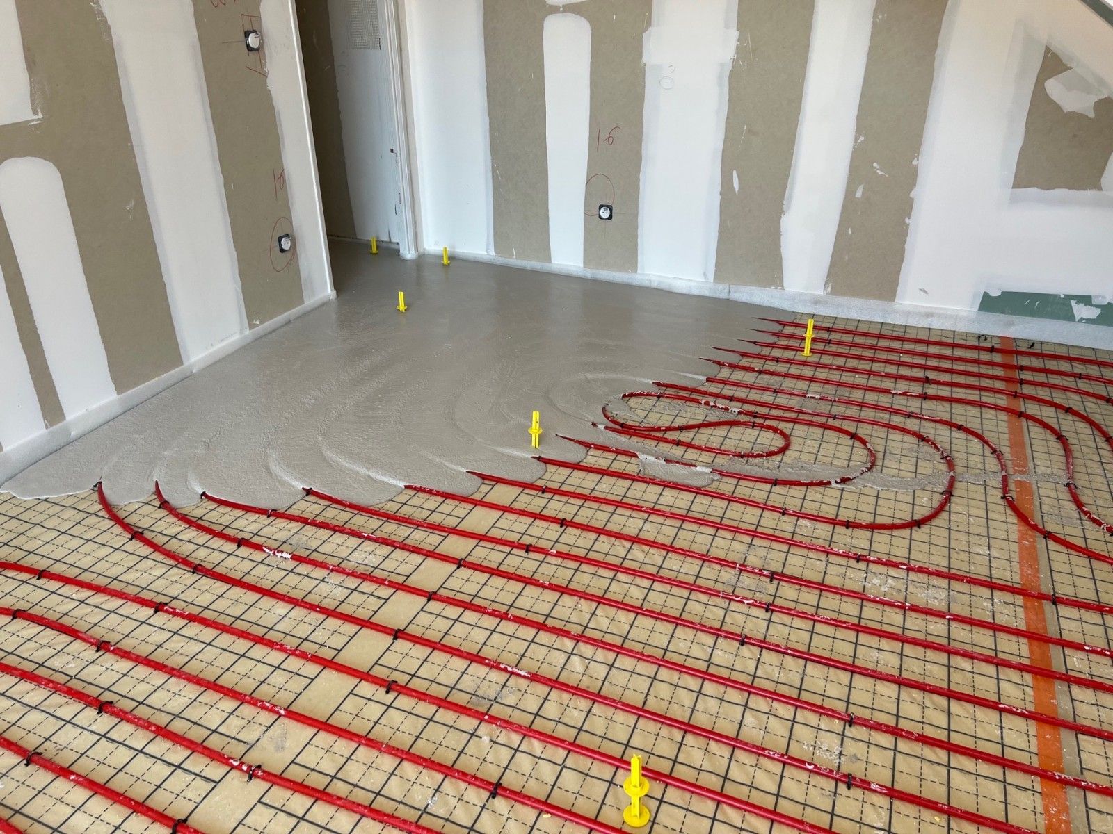 Tuyaux de chauffage au sol radiant installés dans une pièce en construction avec des murs en plaques de plâtre et un sous-plancher non finis.