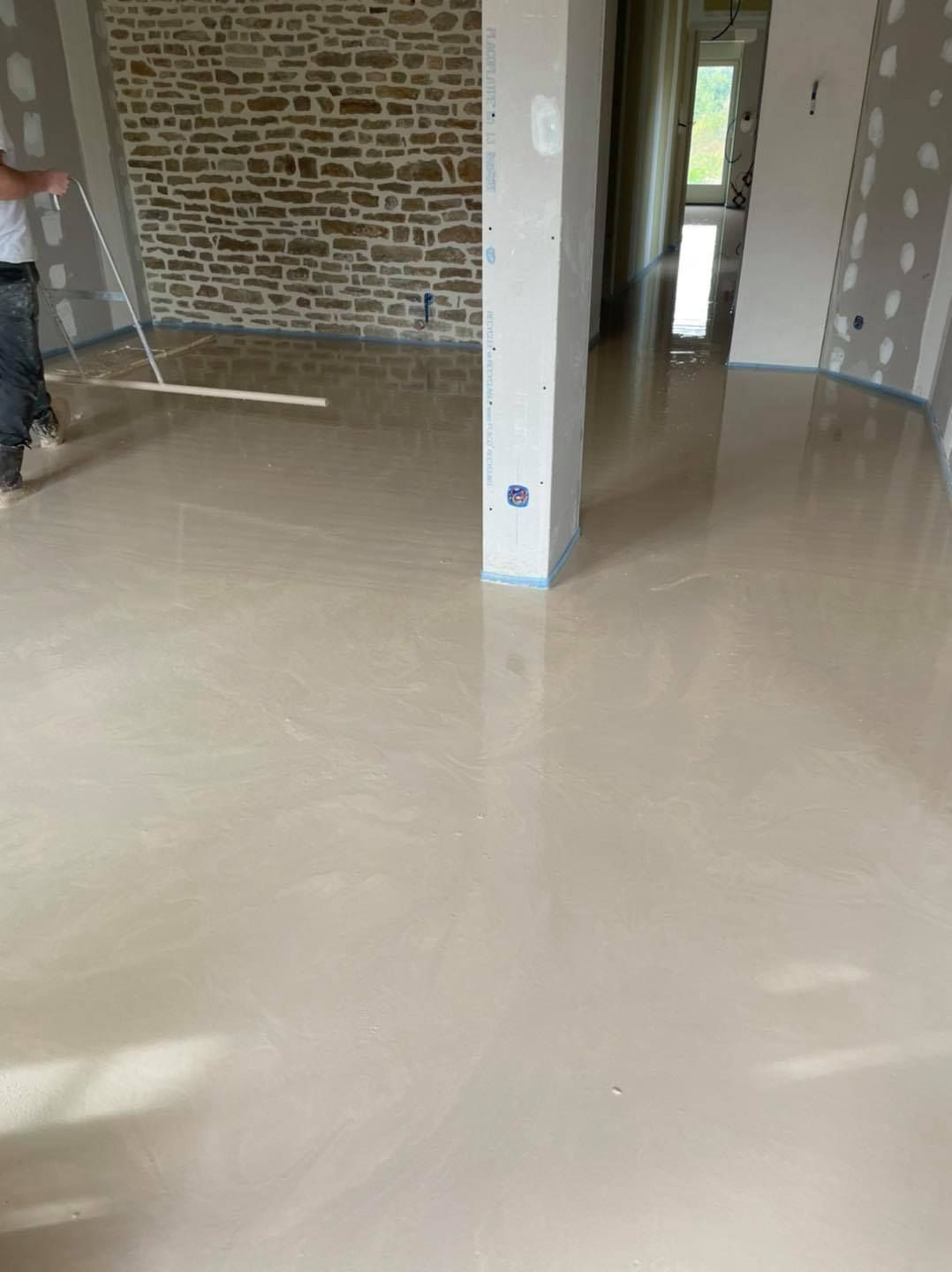 Pièce vide avec sol beige brillant, mur en pierre et une personne qui passe la serpillière à gauche.