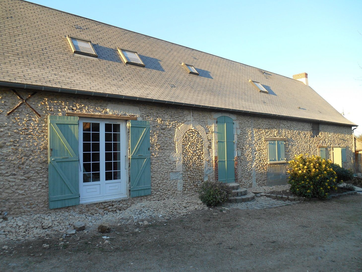 Ferme en pierre avec volets bleus et doubles portes blanches sous un toit de tuiles, vue en plein jour