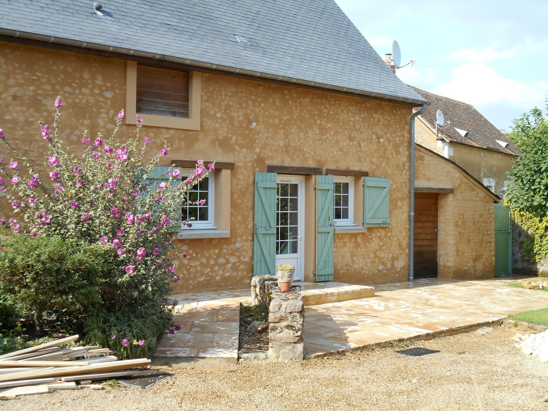 Maison en pierre avec des volets bleus et un buisson à fleurs roses dans la cour avant
