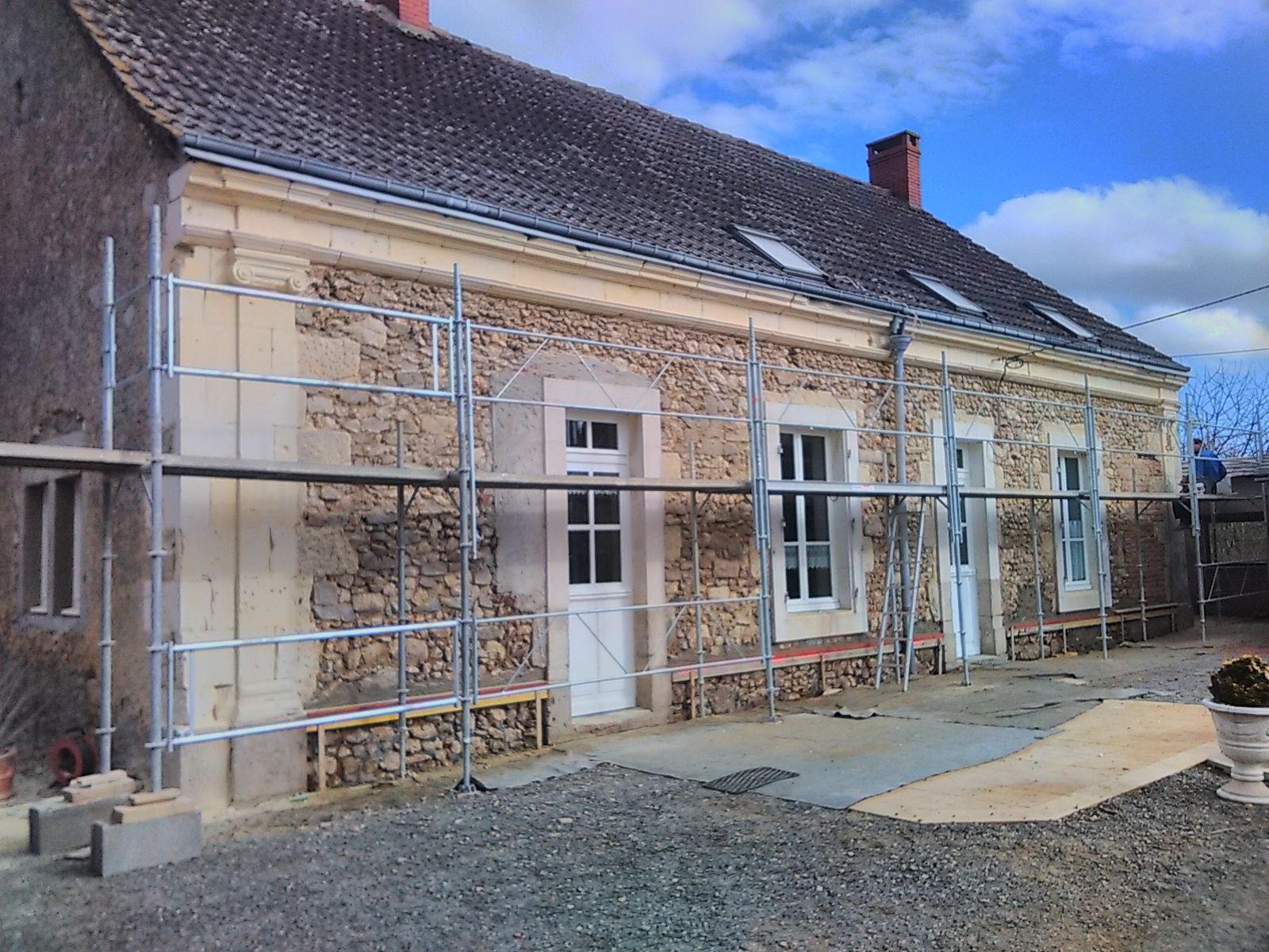 Façade en pierre d'une maison avec des échafaudages autour des fenêtres et de la porte, sous un ciel bleu pendant les travaux de rénovation