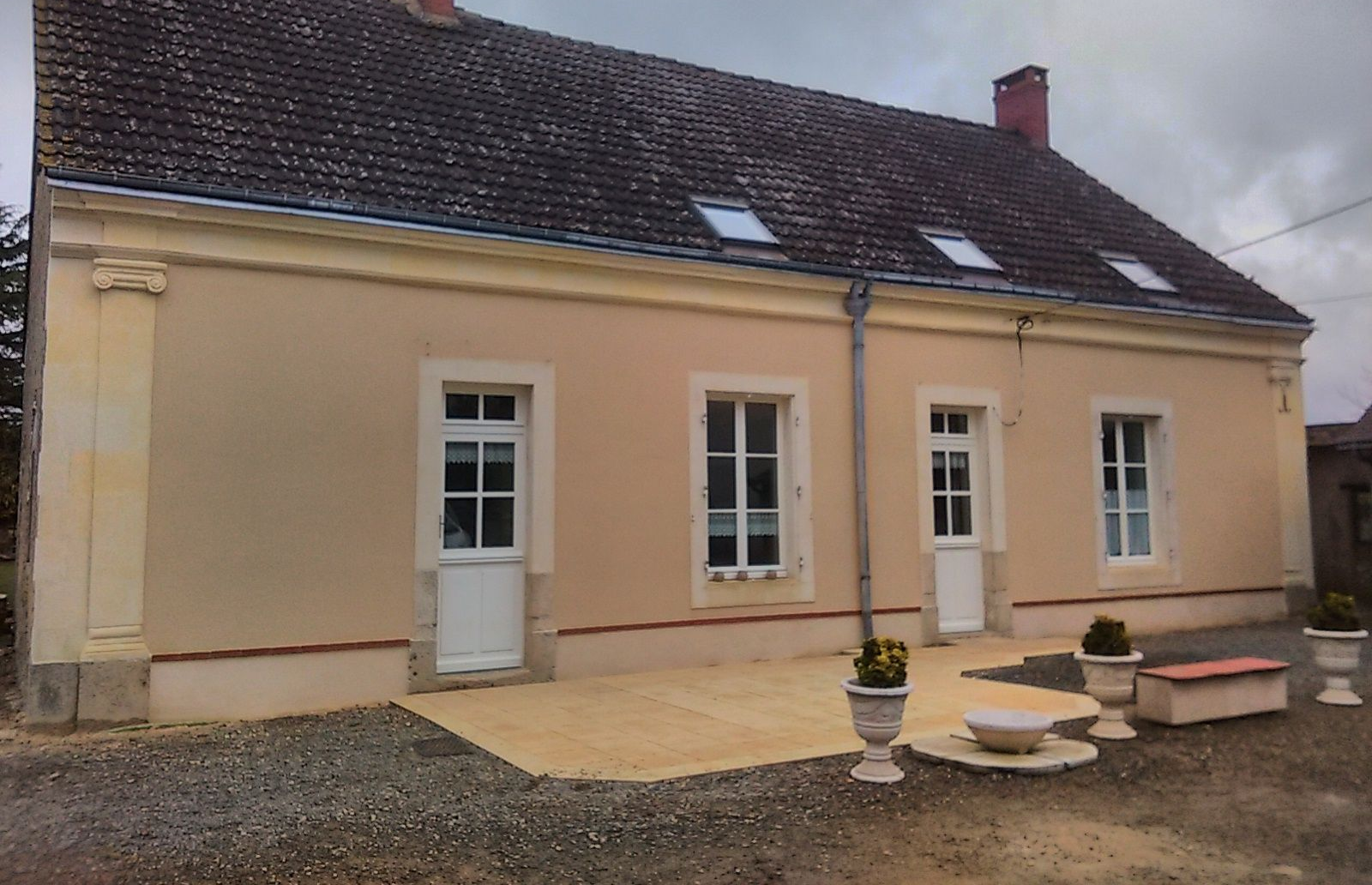 Maisonnette couleur crème avec fenêtres à encadrement blanc, toit rouge et petit patio avec jardinières.
