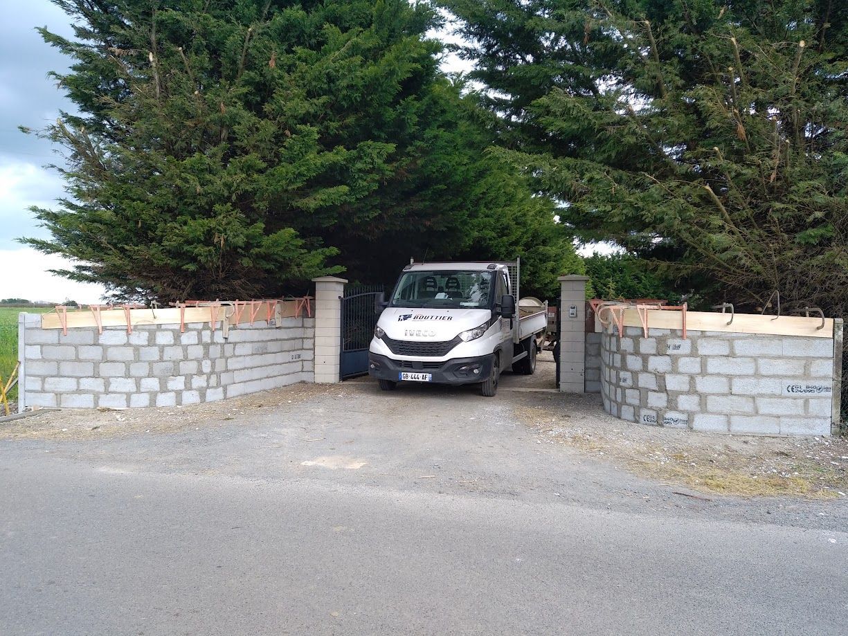 Une camionnette blanche est garée dans une allée privée fermée par un portail, entre des murs de pierre et de grands conifères.