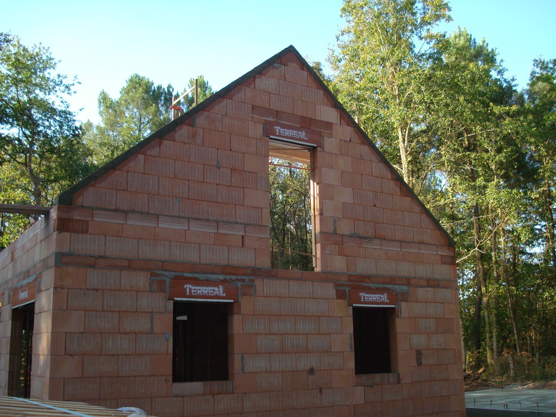 Petite maison en briques rouges en construction, avec deux ouvertures de fenêtres et un toit à pignon élevé, dans une zone boisée.