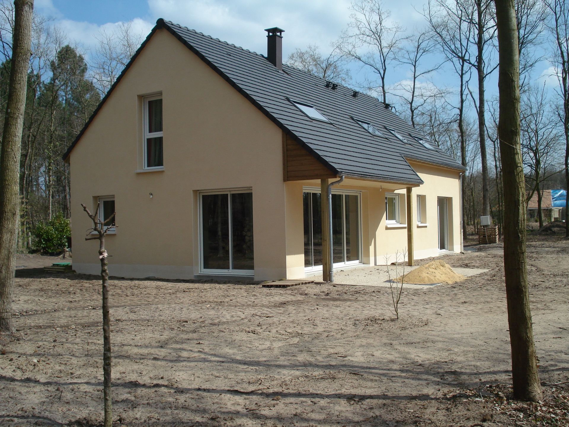 Maison beige au toit gris pentu, située dans une zone boisée, entourée de terrain nu et d'arbres.