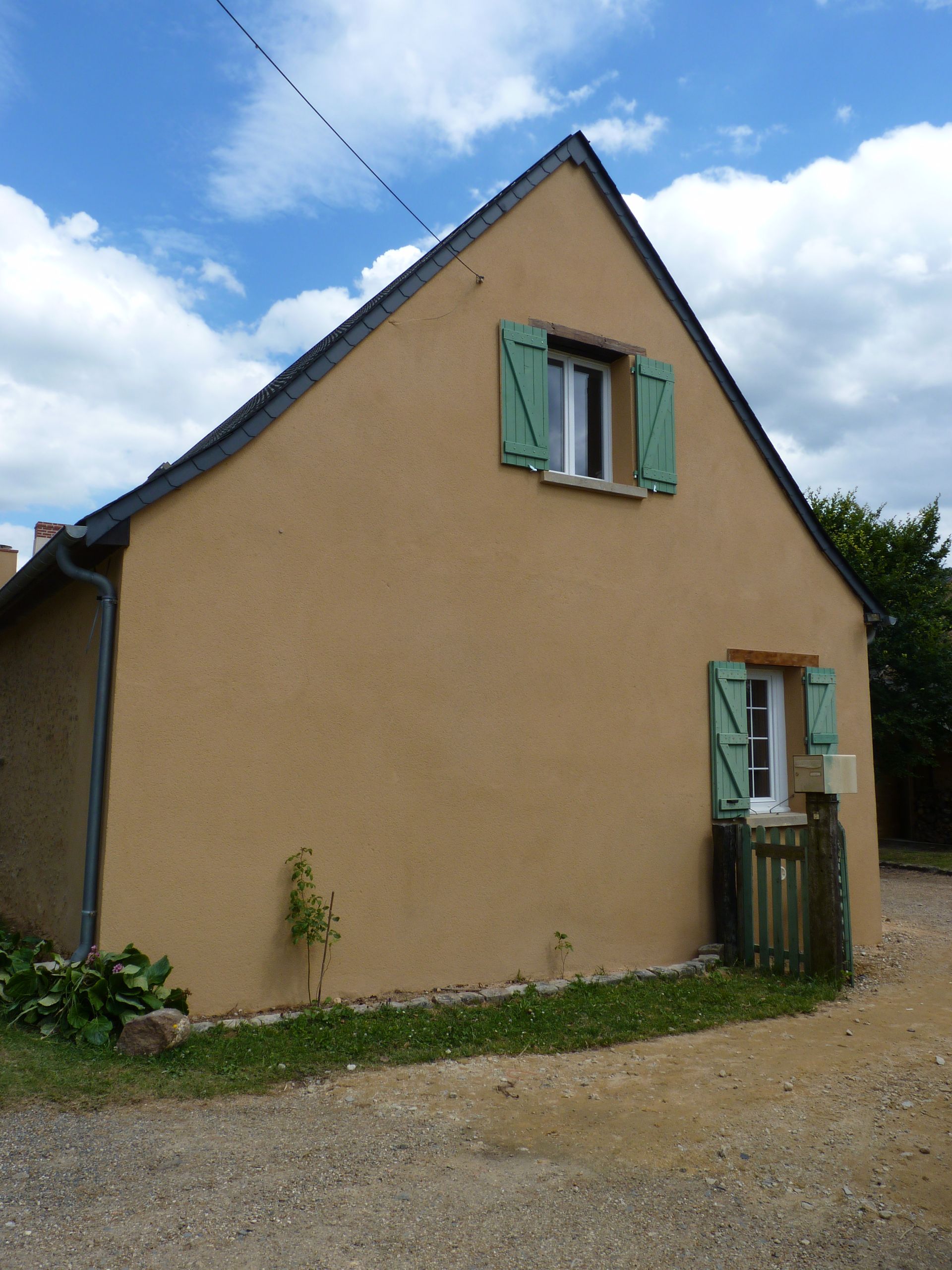 Maison beige aux volets verts sur un chemin de gravier sous un ciel bleu