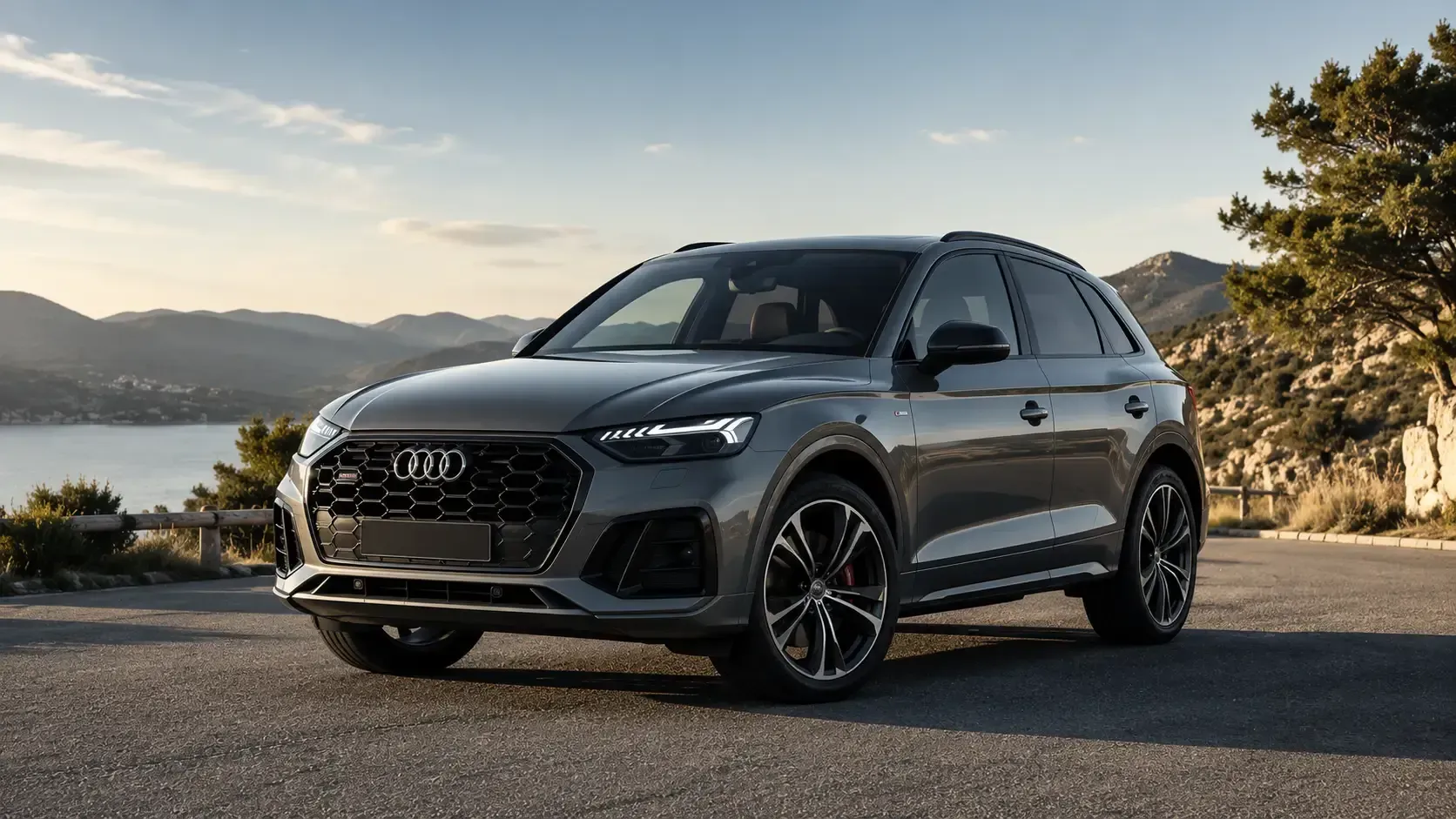 Un SUV Audi gris oscuro estacionado a la orilla de un lago al atardecer, con montañas al fondo.