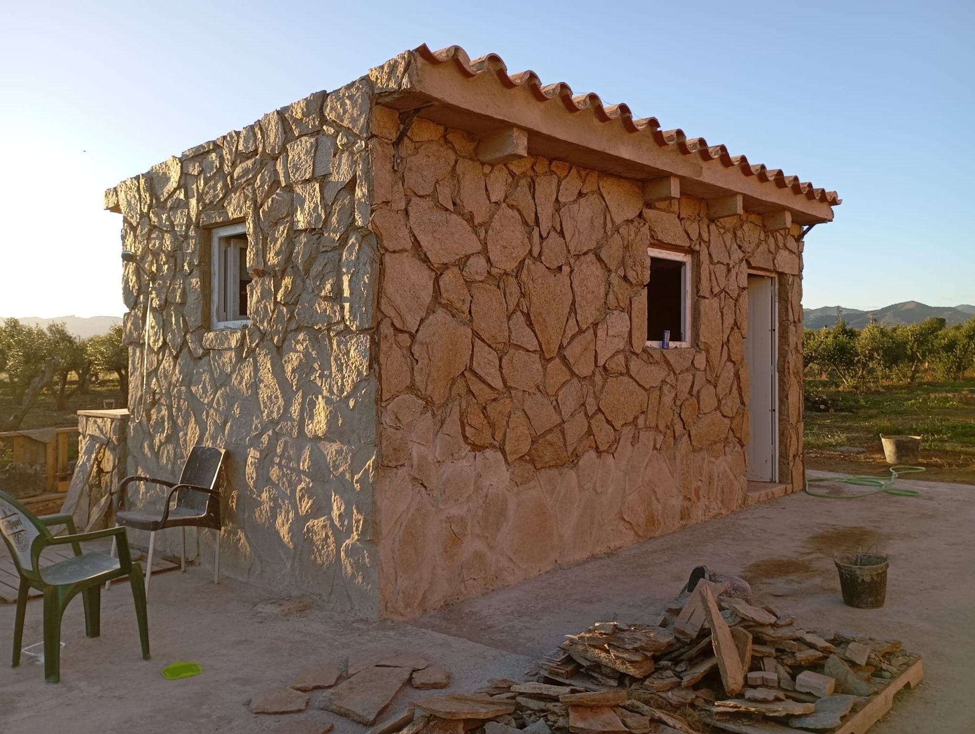 Un pequeño edificio de piedra con una ventana y sillas delante.