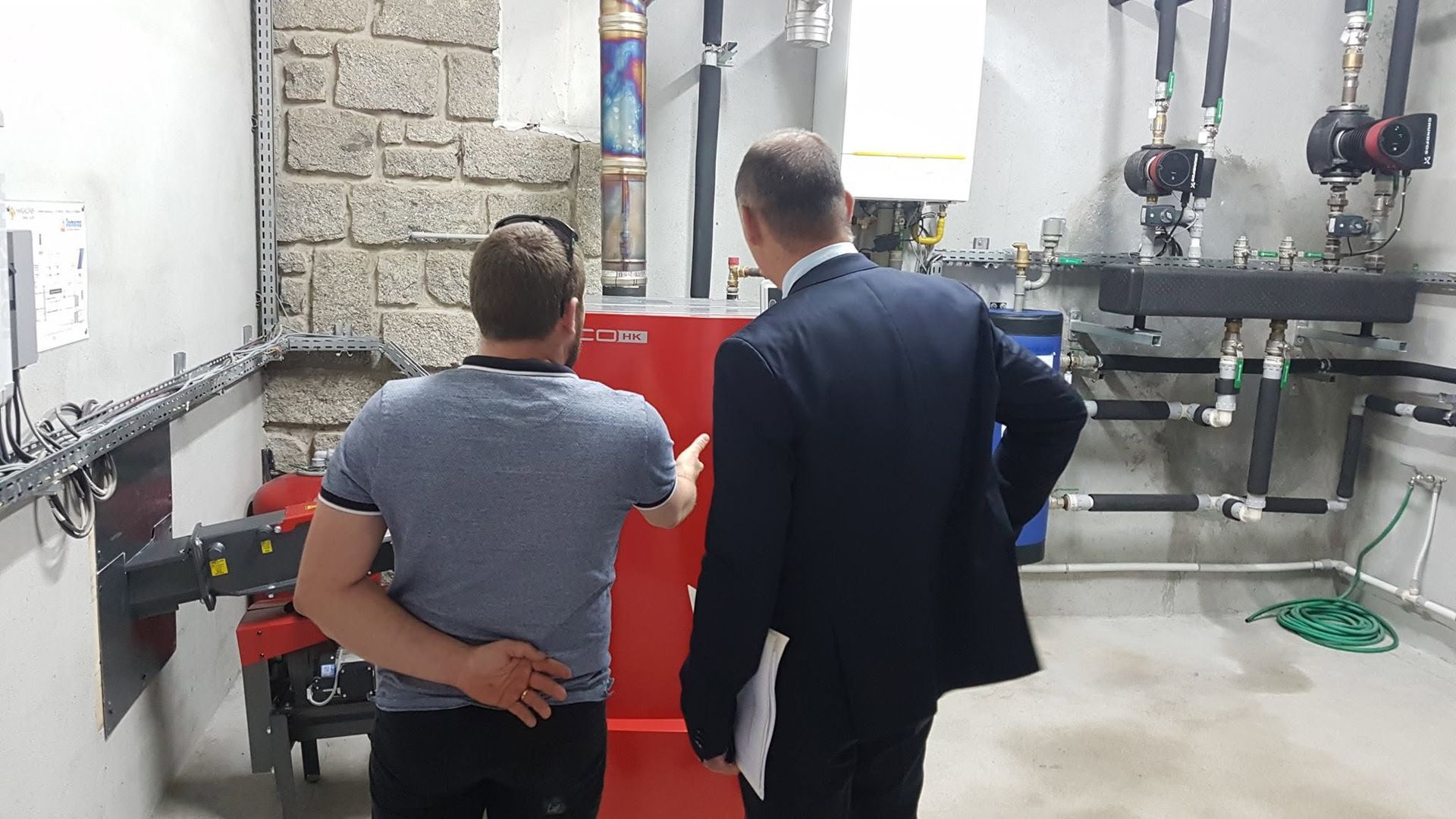 Deux personnes se tiennent dans une chaufferie, observant des équipements industriels rouges et des tuyaux apparents.