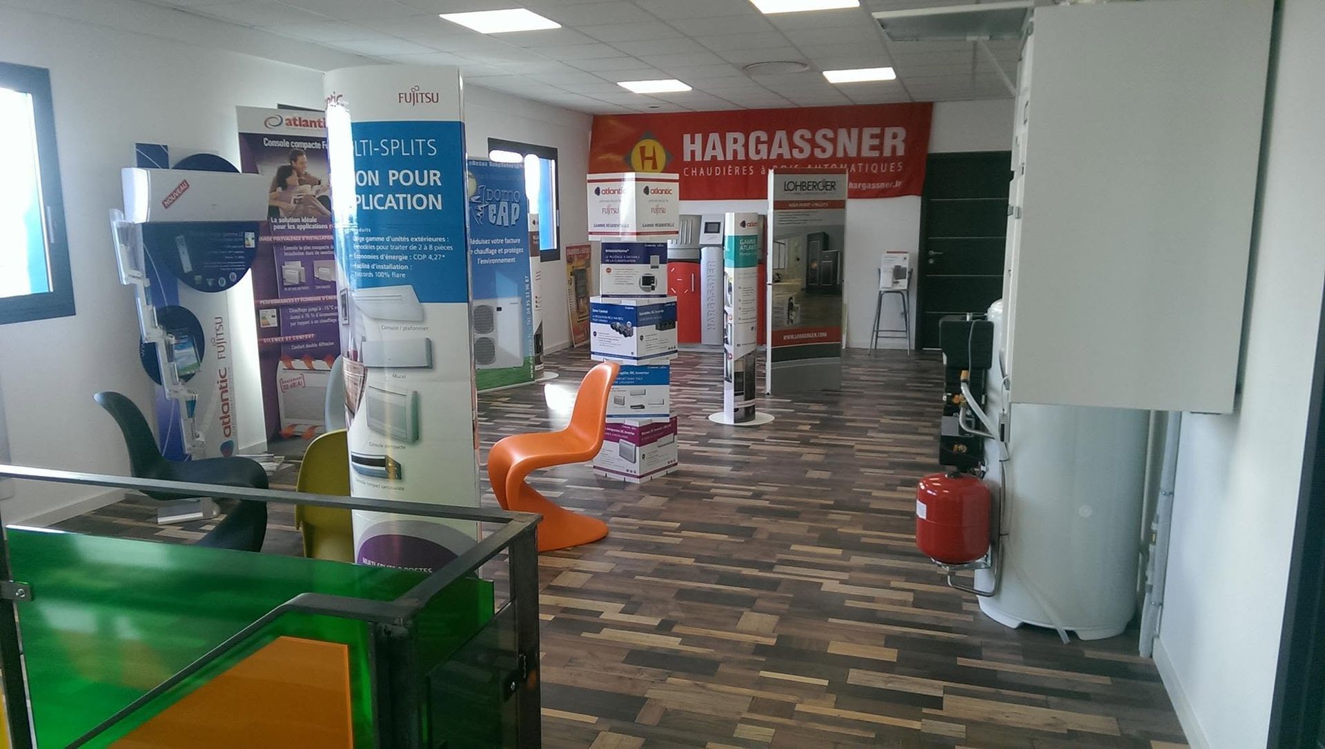 Hall d'entrée intérieur avec sol carrelé, chaises orange, affiches et une cabine Hargassner rouge au fond.
