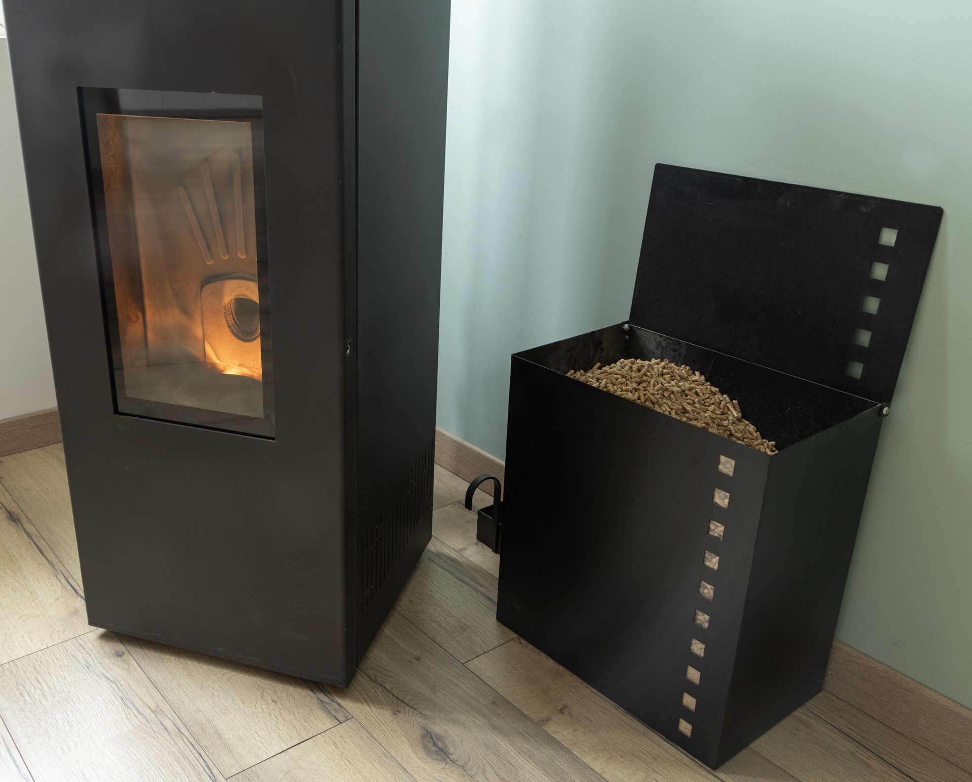 Poêle à granulés noir à côté d'un silo à granulés ouvert, à l'intérieur d'une maison ; les flammes sont visibles à travers la vitre du poêle.
