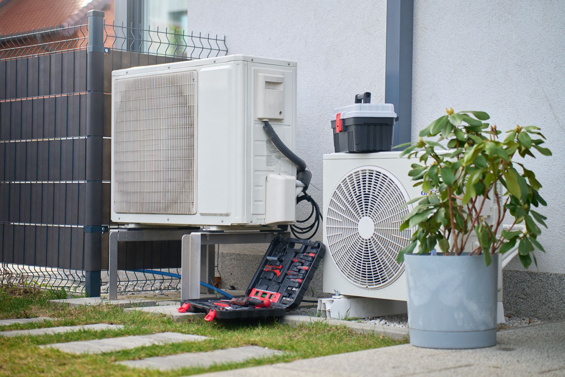 Unités de climatisation extérieures contre un mur, avec des outils au sol et une plante en pot à proximité.