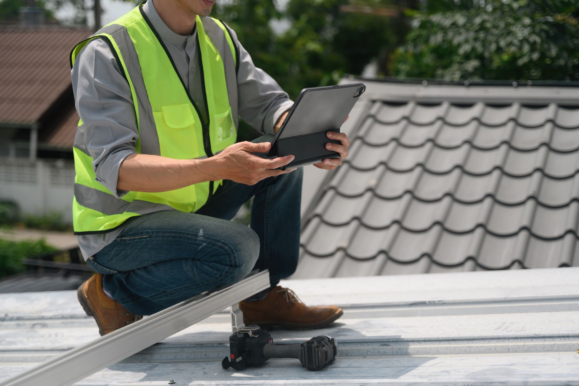 Un ouvrier portant un gilet fluo, agenouillé sur un toit avec une tablette, inspecte les tuiles.