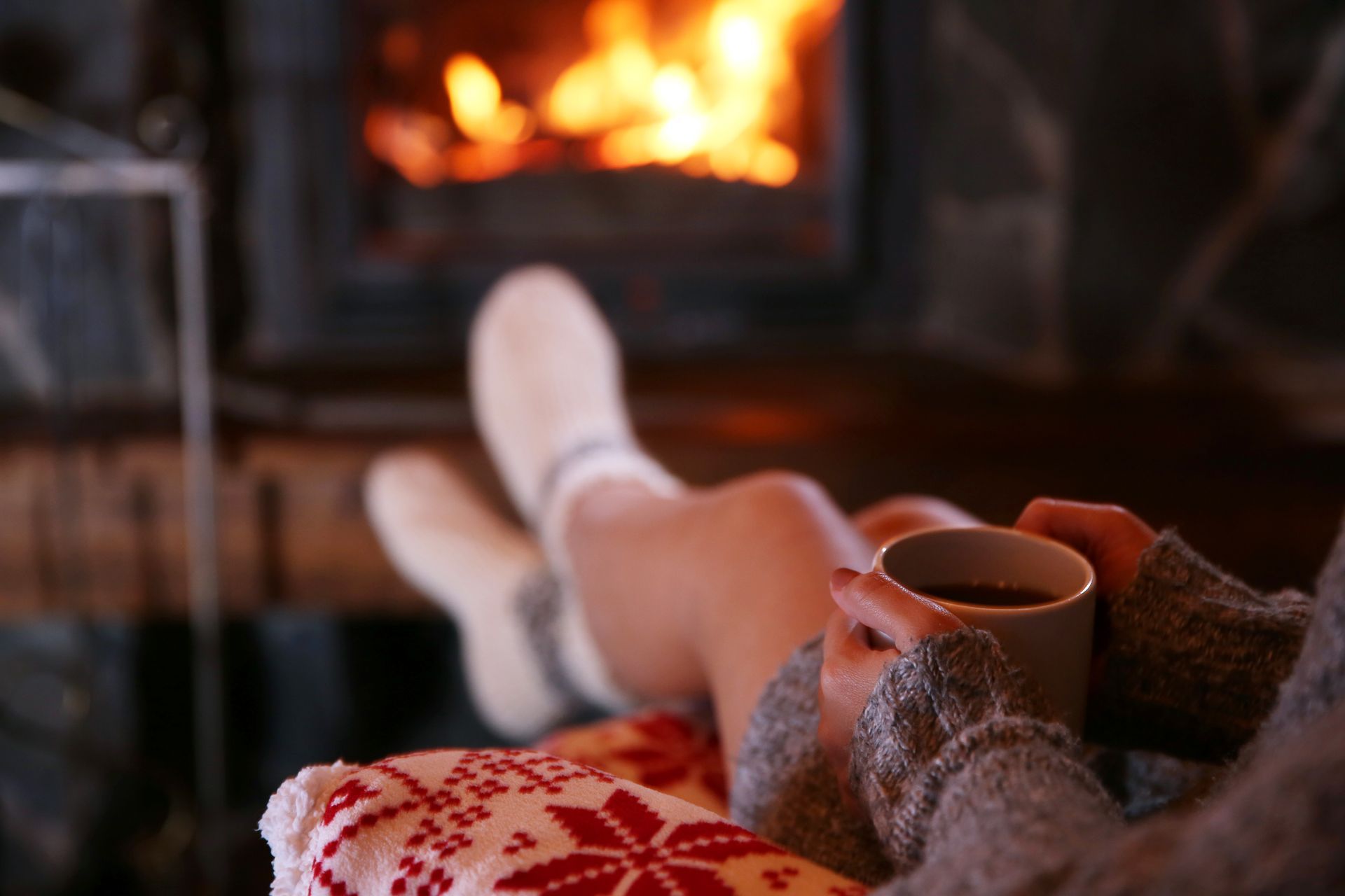 Une personne en chaussettes se détend près d'une cheminée, tenant une tasse et enveloppée dans une couverture à motifs.