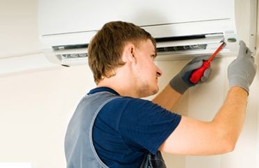 Hombre con guantes grises usando un destornillador en un acondicionador de aire montado en la pared.