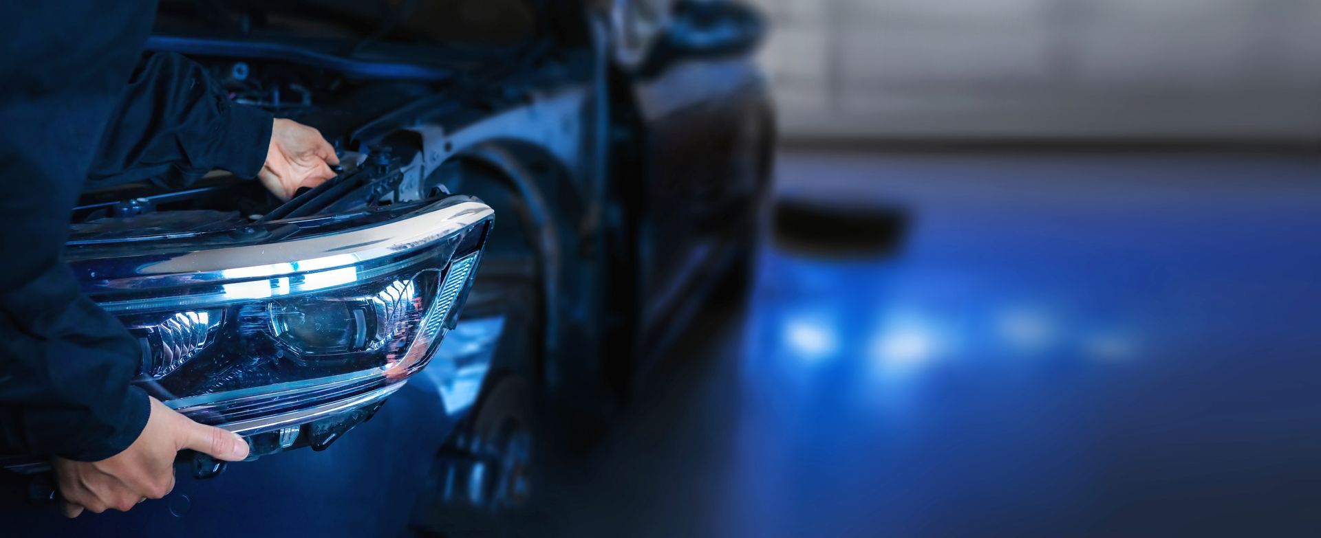 Un mécanicien installe un bloc optique avant sur une voiture de couleur foncée dans un garage éclairé en bleu.