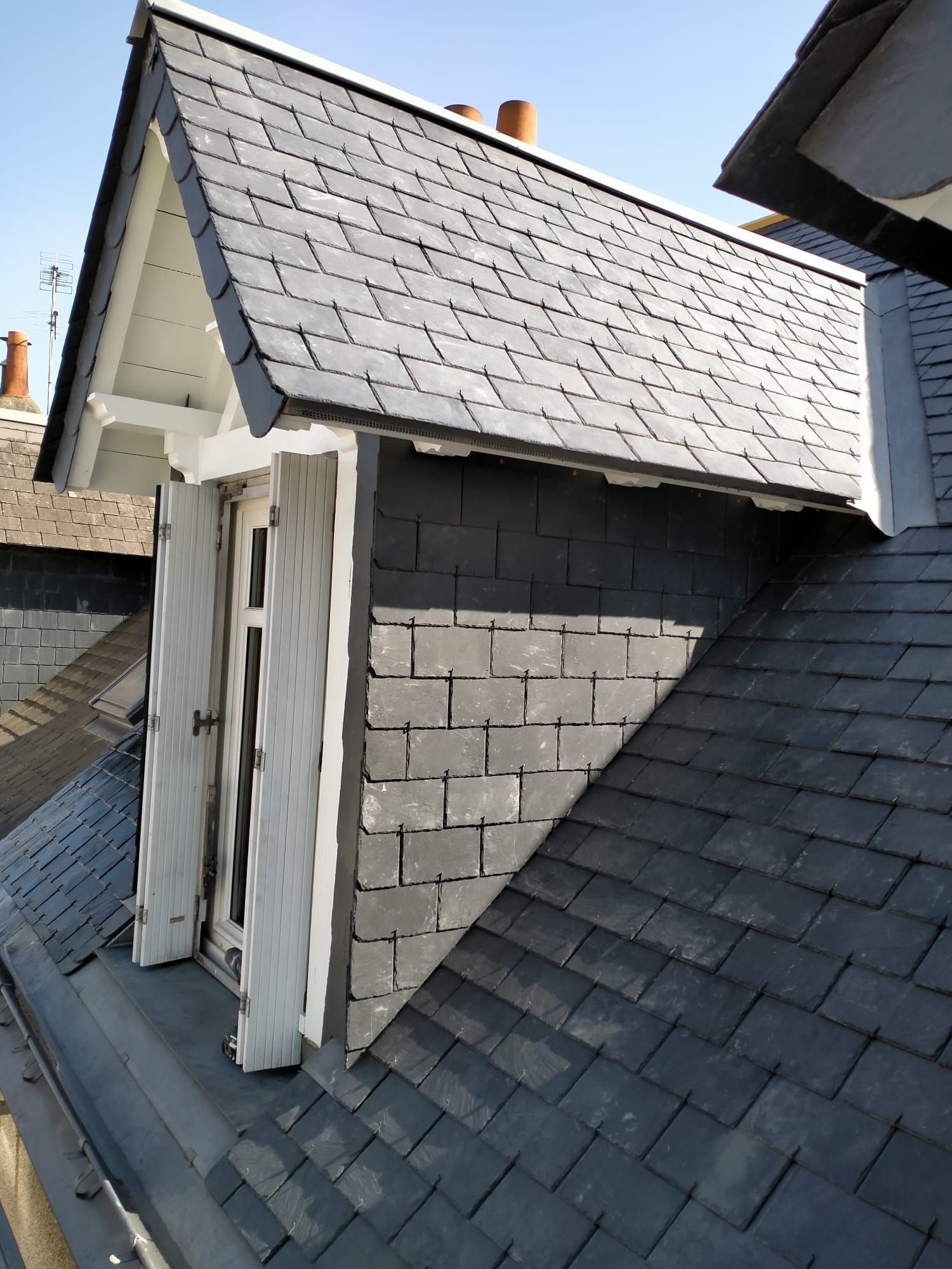 Gros plan sur une lucarne de toit avec volets blancs et toiture en ardoise