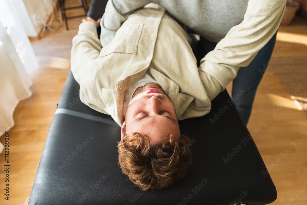 Eine Person liegt kopfüber auf einer schwarzen Massageliege und trägt ein beiges Hemd in einem hellen Raum.