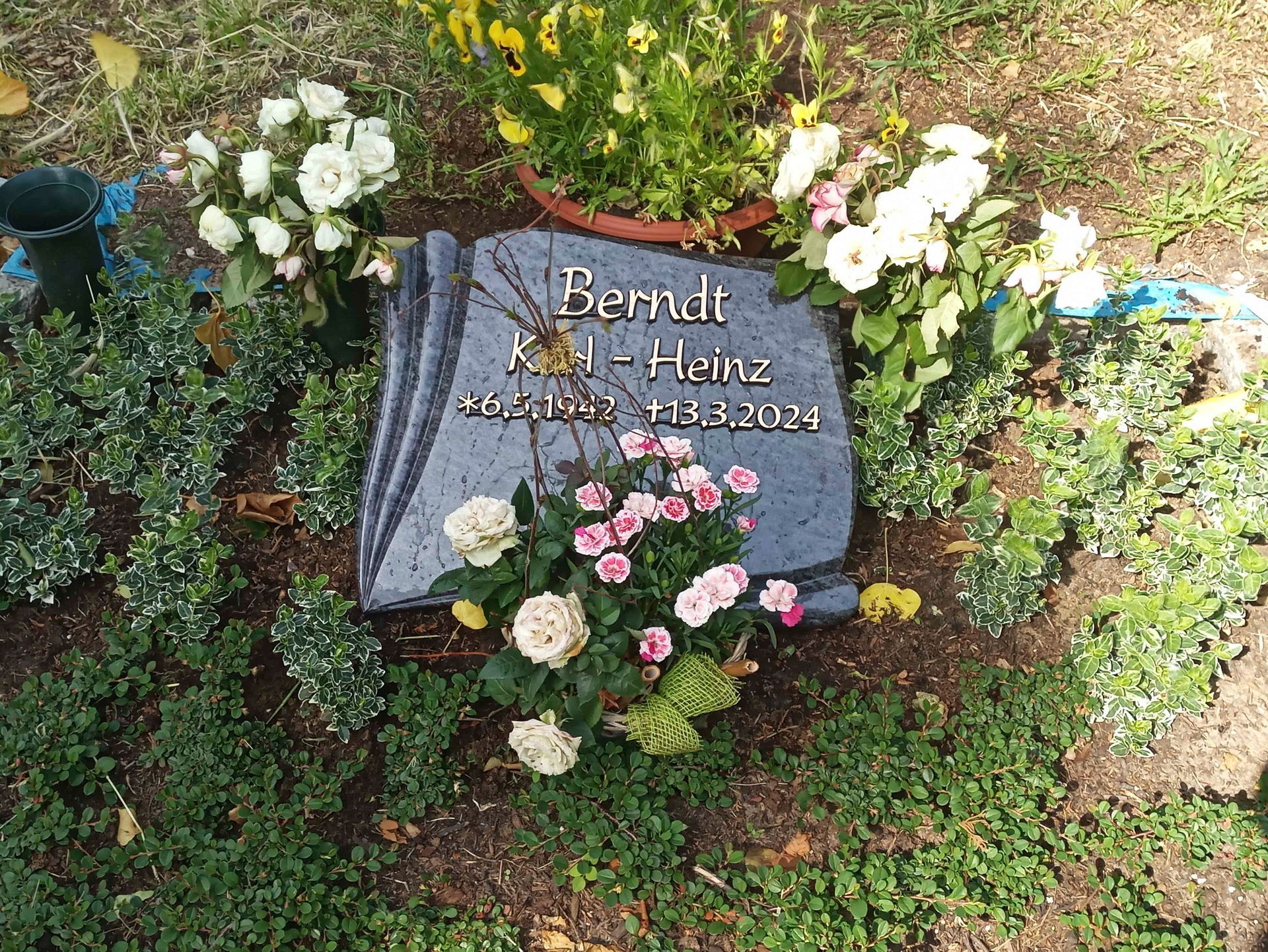 Ein Grabstein in Form eines Buches ist von Blumen umgeben.