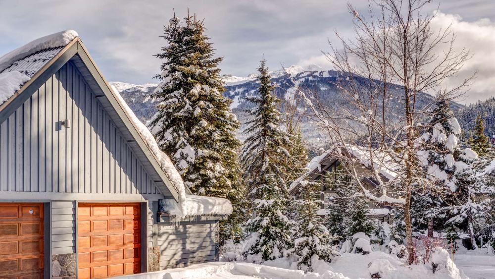 A snow-covered garage with wooden doors sits near evergreen trees and mountains under a cloudy sky.