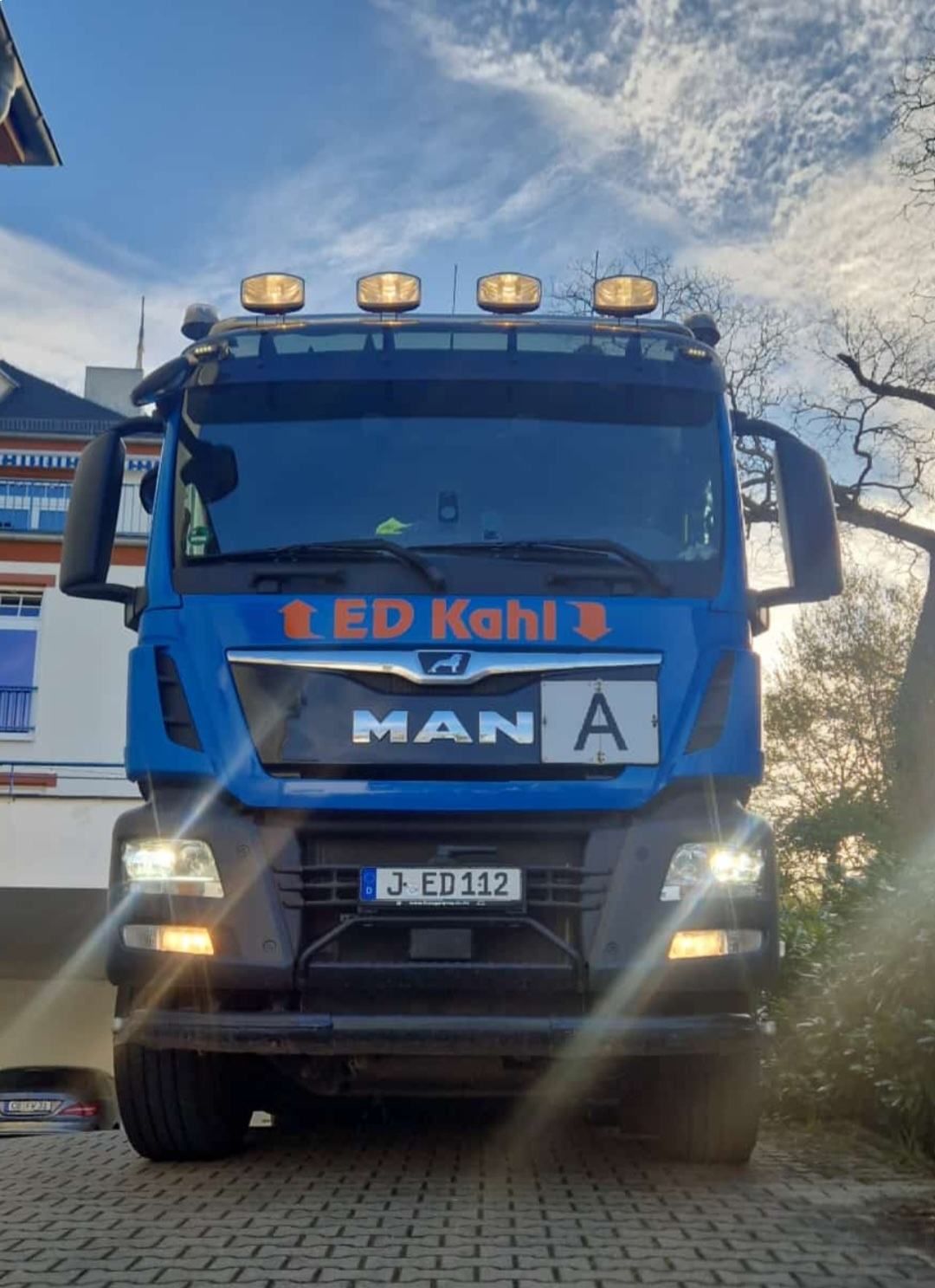 Ein blauer MAN-Lkw mit eingeschalteten Scheinwerfern fährt auf einer Straße unter bewölktem Himmel