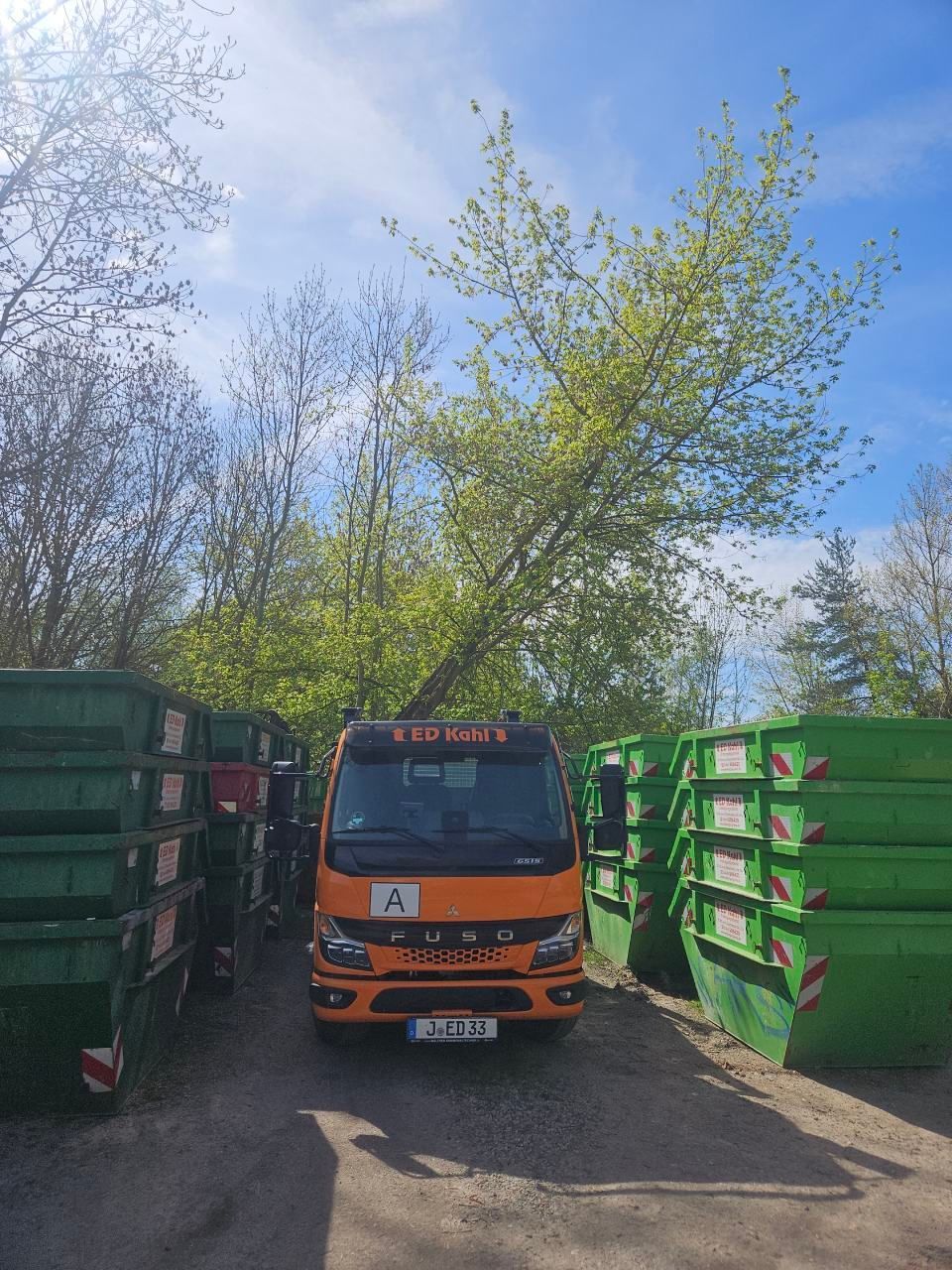 Ein orangefarbener Lastwagen zwischen gestapelten grünen Mülltonnen auf einem schmalen Feldweg unter Bäumen und blauem Himmel