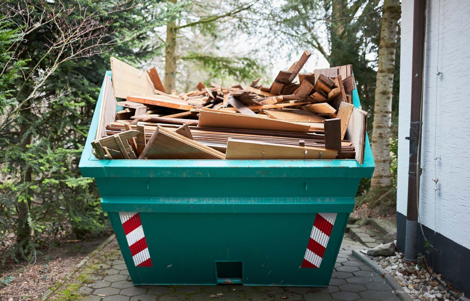 Ein Arbeiter mit Handschuhen reinigt im Freien einen großen grünen Metallbehälter mit einem Abzieher.