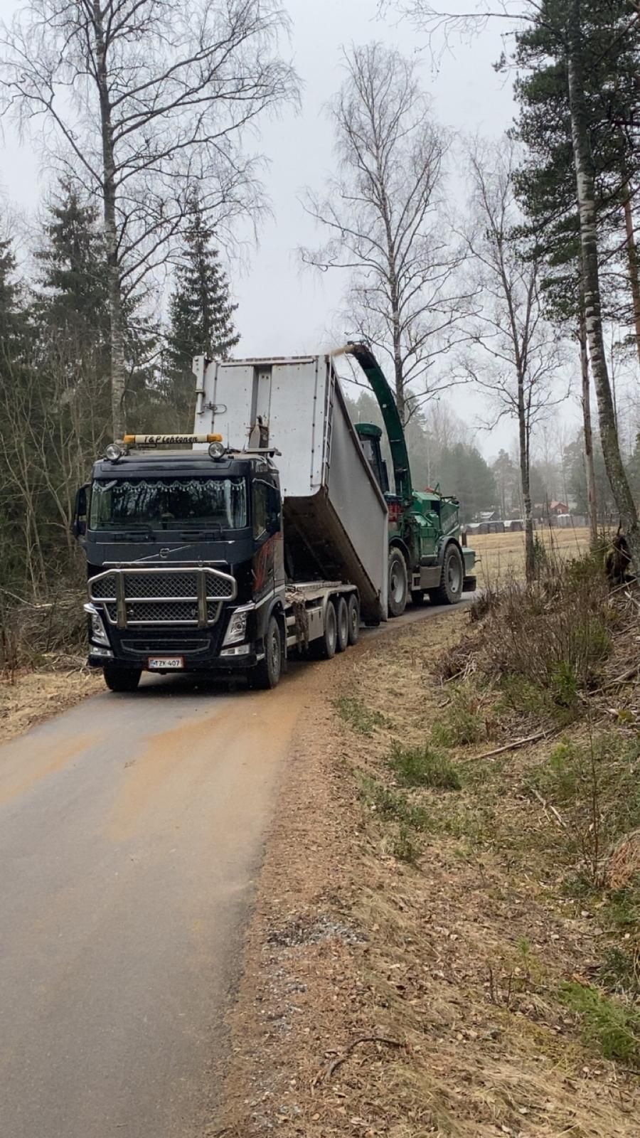 Kuorma-auto purkaa suurta valkoista konttia hiekkatiellä metsäalueella.