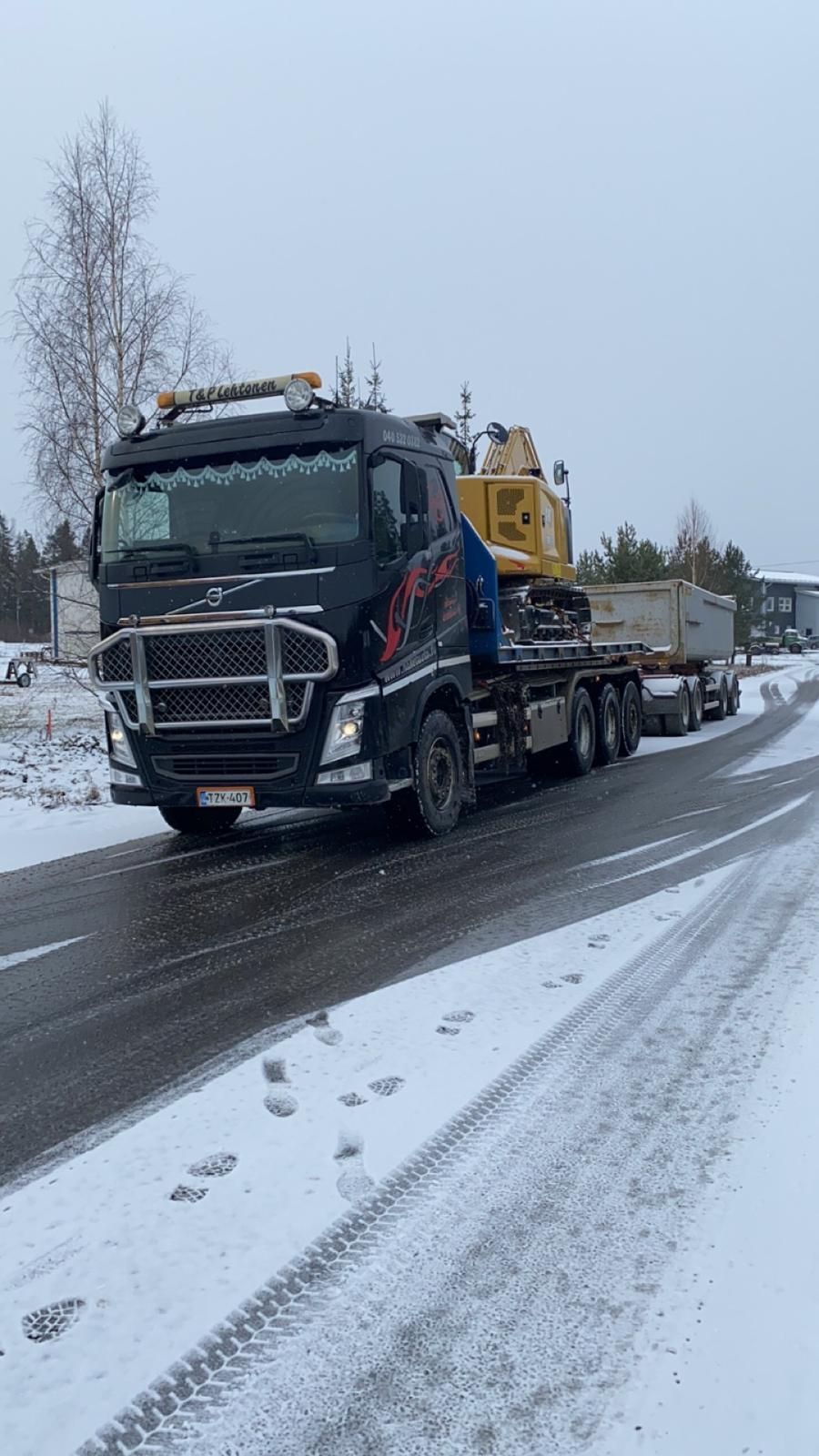 Musta rekka-auto vetää raskaita koneita lavetilla lumipeitteisellä tiellä.