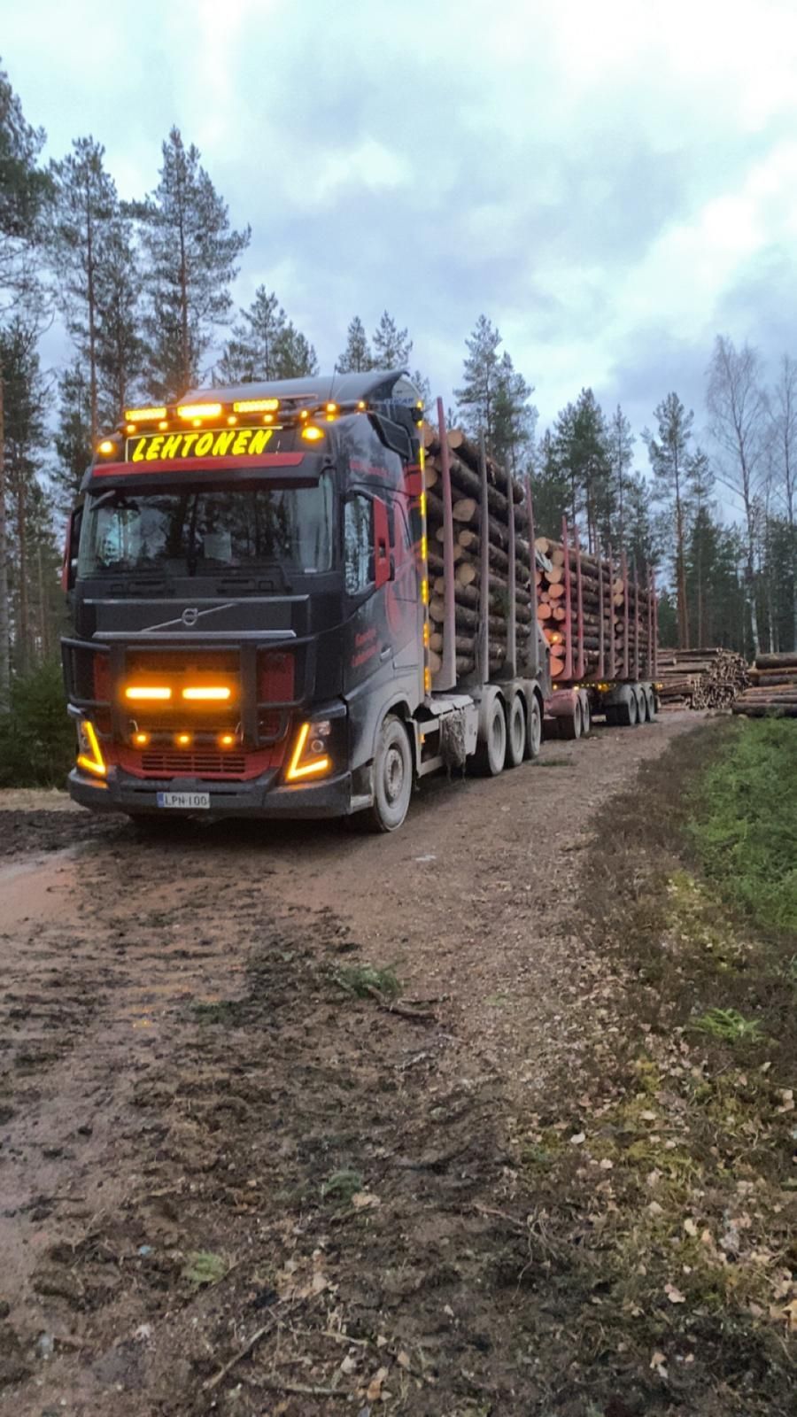 Musta tukkiauto lastattuna tukkeilla mutaisella metsätiellä, pilvinen taivas.